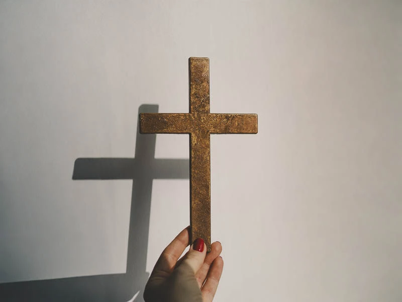 Dłoń trzymająca prosty drewniany krzyż rzucający cień na białą ścianę, symbolizujący istotę wiary chrześcijańskiej i zbawienie przez krzyż