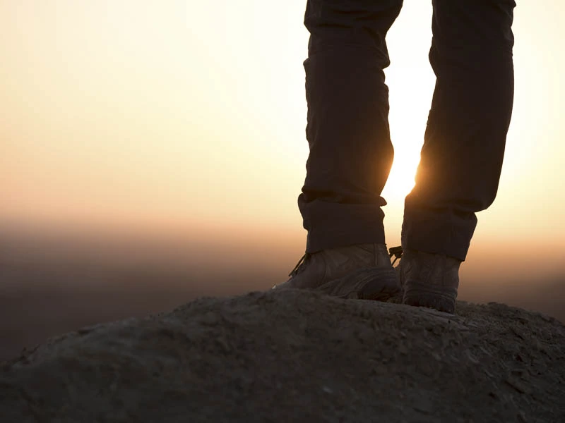 Sylwetka nóg osoby stojącej na skalistym wzniesieniu na tle zachodzącego słońca, symbolizująca nowy początek, podróż wiary i pierwsze kroki w życiu chrześcijańskim