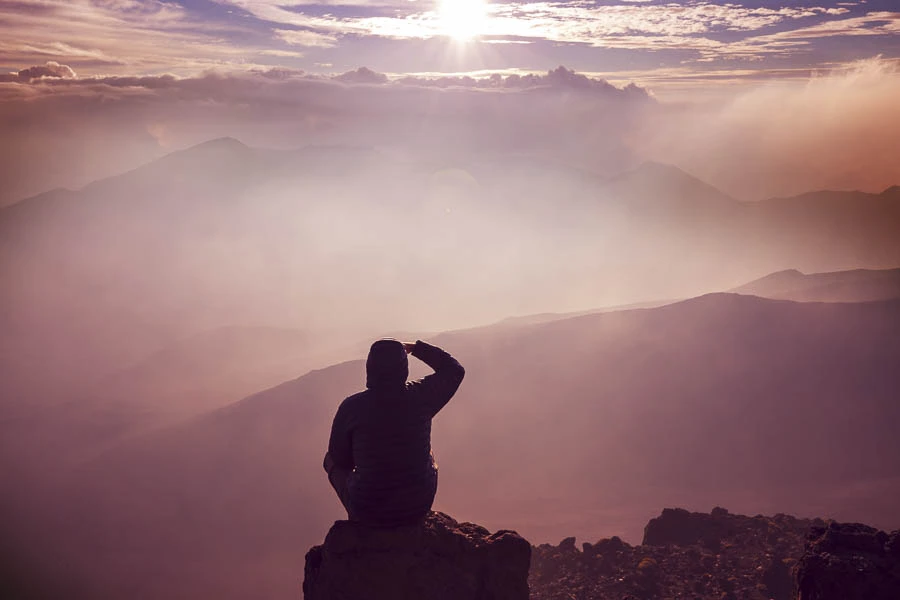 Sylwetka osoby siedzącej na szczycie góry, patrzącej na majestatyczny wschód słońca ponad mglistymi górami, symbolizująca poszukiwanie sensu życia i duchową refleksję
